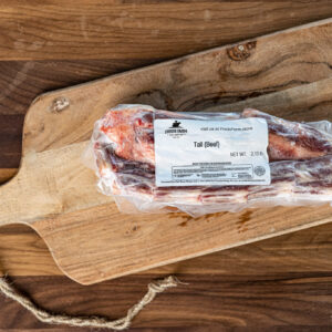 Beef tail package in plastic wrap on a wooden cutting board; label reads Tail (Beef) and shows Fred's Farm branding.