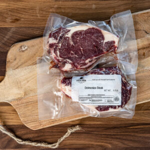 Vacuum-sealed Delmonico steak on a wooden cutting board, label reads 'Delmonico Steak' from Fred's Farm; raw beef.
