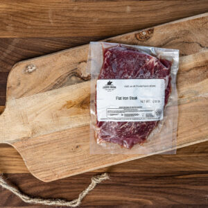 Vacuum-sealed flat iron steak on a rustic wooden cutting board with a labeled package standing upright on the board surface.