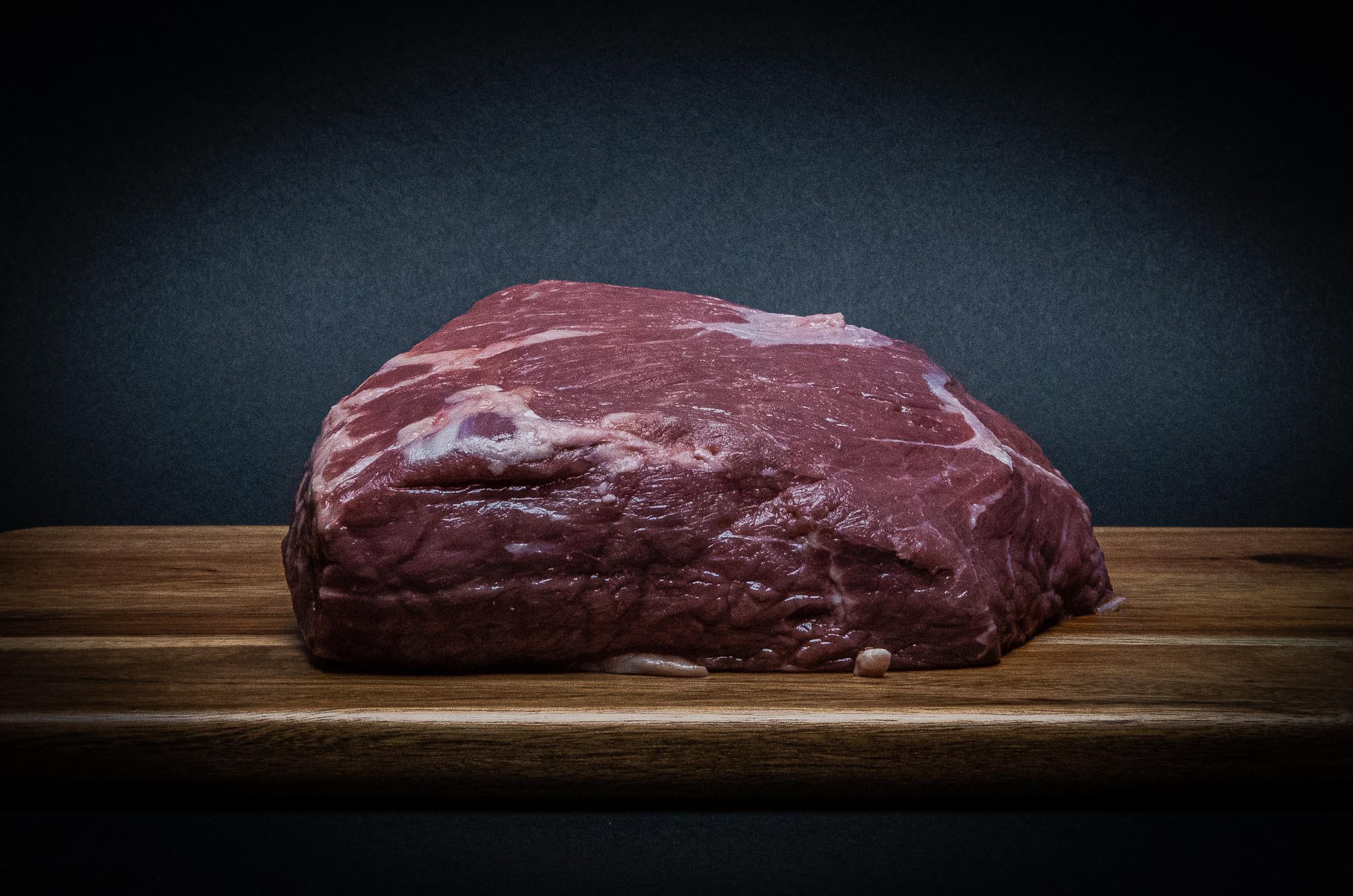 Thick raw beef steak with visible marbling on a wooden cutting board against a dark background.