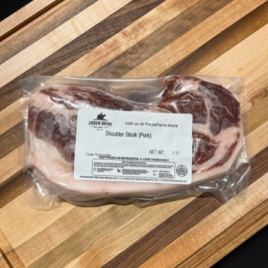 Vacuum-sealed bag of pork shoulder steak on a wooden cutting board; label reads 'Shoulder Steak (Pork)' from Fred's Farm.