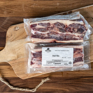 Vacuum-sealed package of short ribs on a wooden cutting board, freezer-safe label visible beyond the plastic wrap (frozen meat).