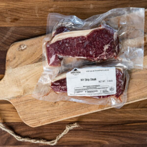 Vacuum-sealed raw NY strip steak on a wooden cutting board with a label reading Fred's Farm.