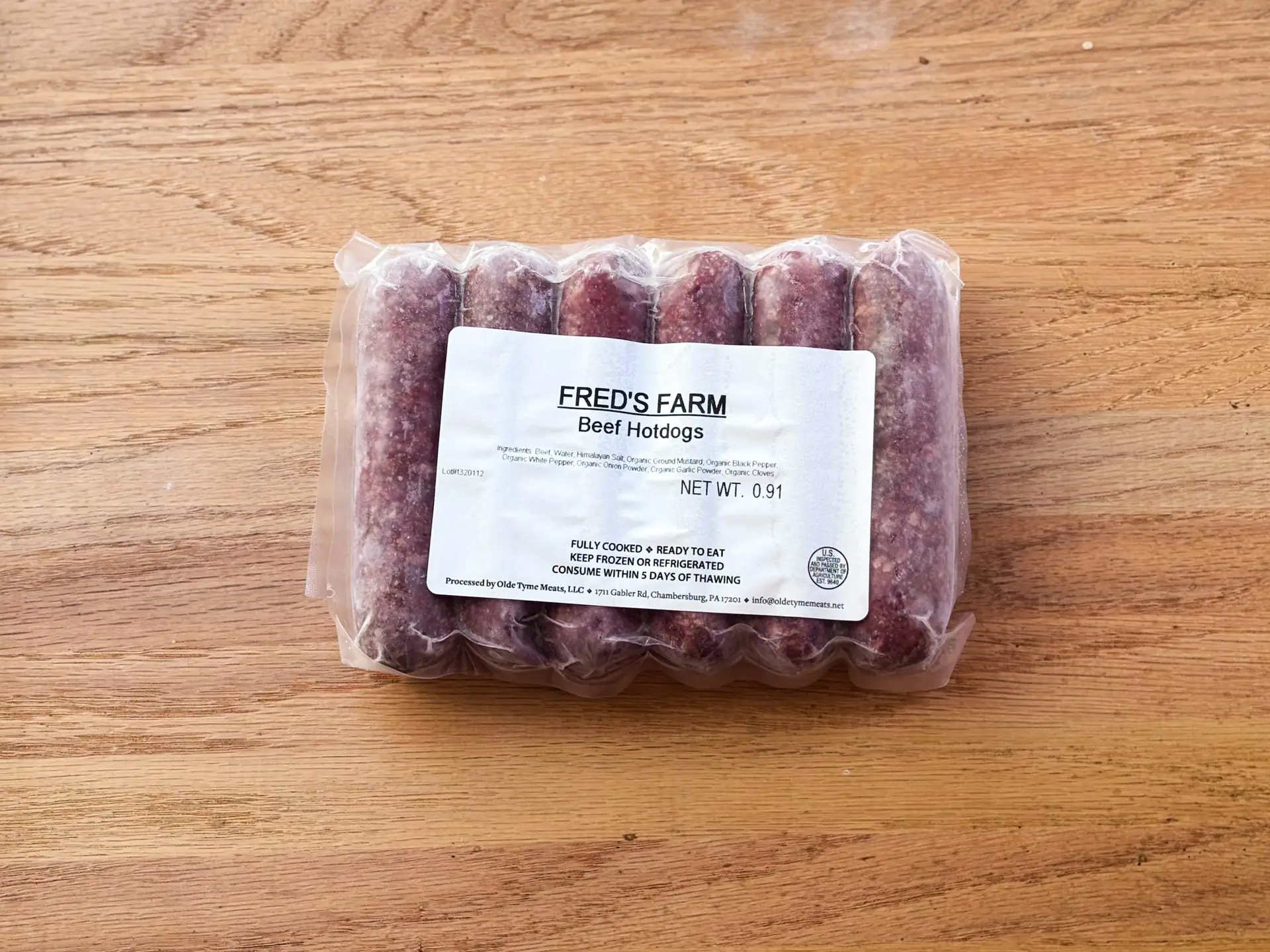 Pack of beef hotdogs in clear plastic packaging on a wooden surface, label reads 'FRED'S FARM'