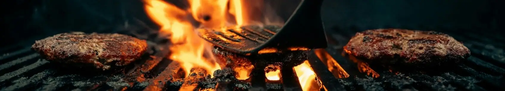 Beef burger patties grilling on a charcoal barbecue with a spatula resting on one patty and flames underneath.